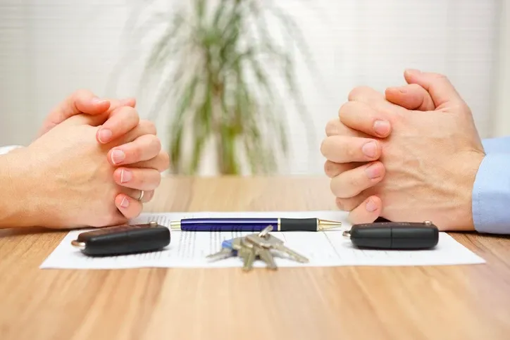 Two pairs of folded hands facing each other across a table with documents and keys between them.